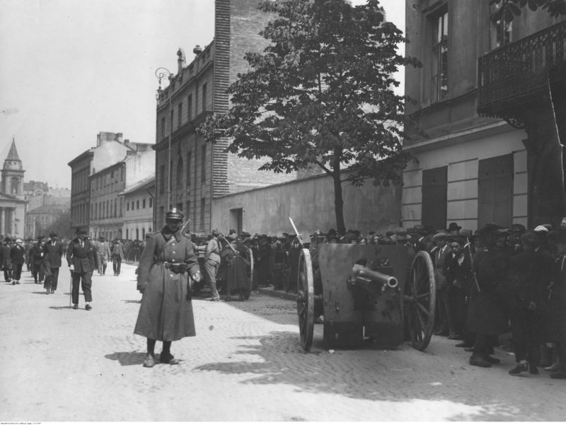 Widok na brukowaną ulicę. Na jej skraju stoją dwa działa polowe obsadzone przez żołnierzy i zwrocone w dwie strony. Na chodniku obok tłum gapiów.