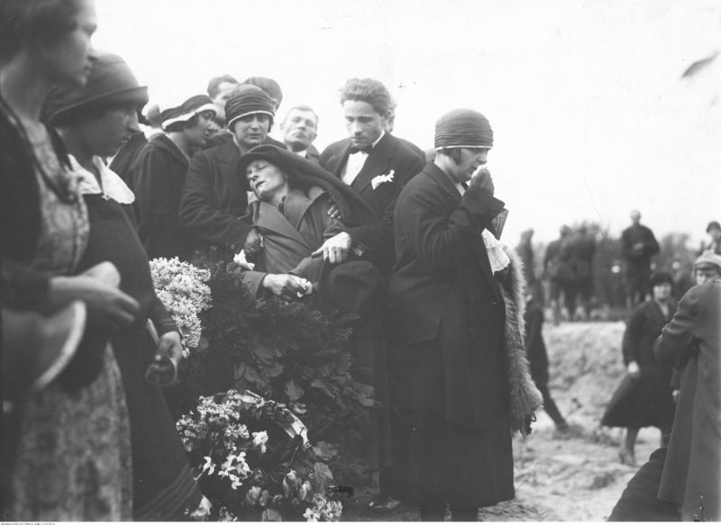 Grupa żałobników stojąca nad grobem z wieńcami kwiatów. Na wprost fotografa mężczyzna i kobieta podtrzymują omdlałą kobietę.