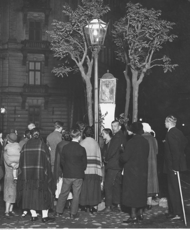 Noc. Na słupie obok ulicznej latarni widoczna niewielka kapliczka, wokół której stoi grupa ludzi zwrócona do fotografa plecami. W tle dwa drzewa i kamienica.