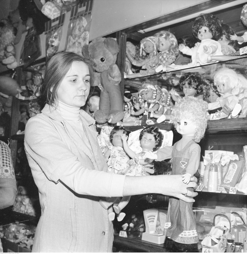 Ekspedientka przy półkach z zabawkami, trzyma w wyciągniętych rękach dużą lalkę.