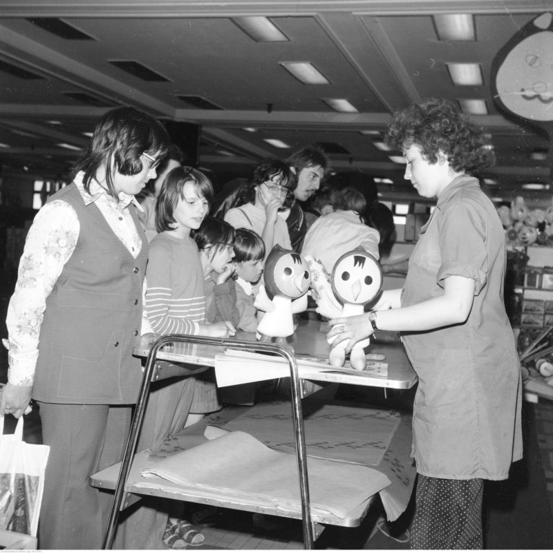 Stoisko z zabawkami. Ekspedientka prezentuje klientom dmuchane zabawki. Widoczne dzieci.