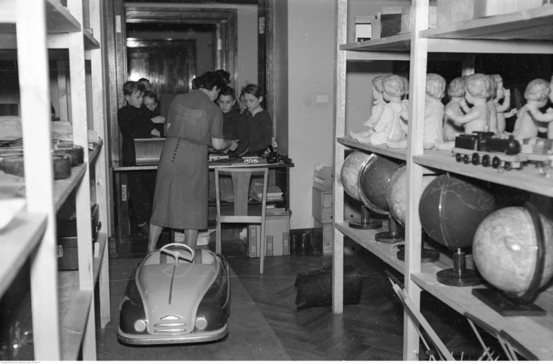 Na półkach widoczne lalki oraz globusy. Na podłodze stoi samochód na pedały.