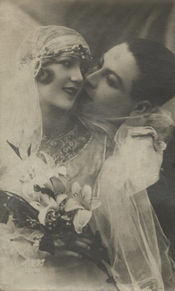 Para młoda w strojach ślubnych ukazana w półpostaci. Ona trzyma bukiet z lilii, on zbliża usta do jej policzka.