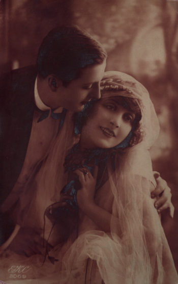 Para w strojach ślubnych, mężczyzna pochylający się i obejmujący siedzącą kobietę. Zdjęcie o wyraźnym czerwonym zabarwieniu.