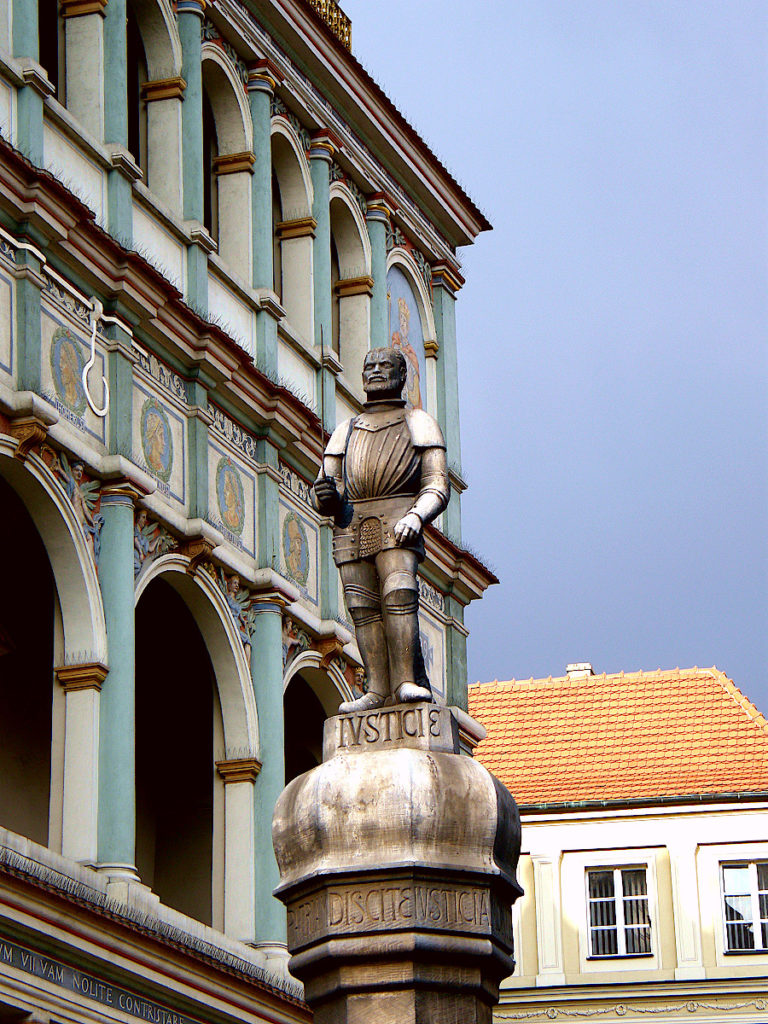 Jeden z napisów na poznańskim pręgierzu głosi: „Wybudowano ten posąg z fatałaszków kucharek roku Pańskiego 1535” (fot. Kwolana, CC BY-SA 4.0) Czarna figurka rycerza wieńcząca poznański pręgierz. W tle widoczny fragment fasady ratusza.
