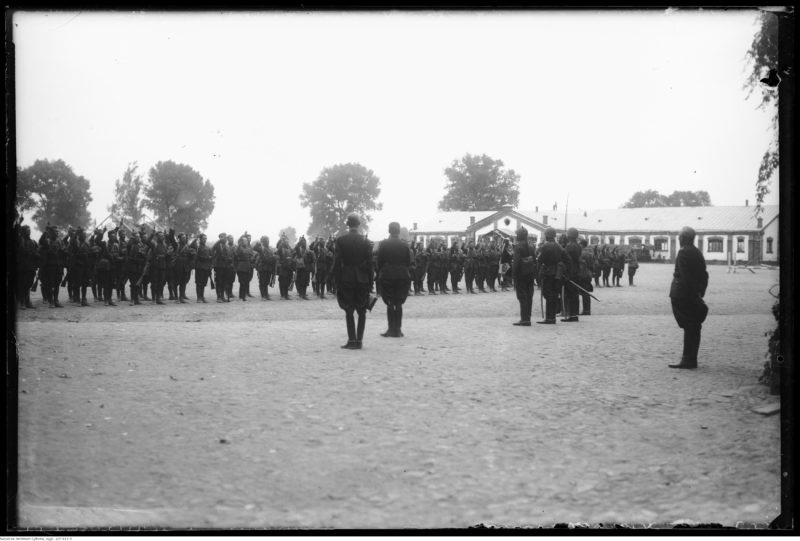 Na obszernym ziemnym placu żołnierze w dwuszeregu z prawymi rękoma uniesieonymi do przysięgi. Naprzeciw nich oficerowie oraz poczet sztandarowy.