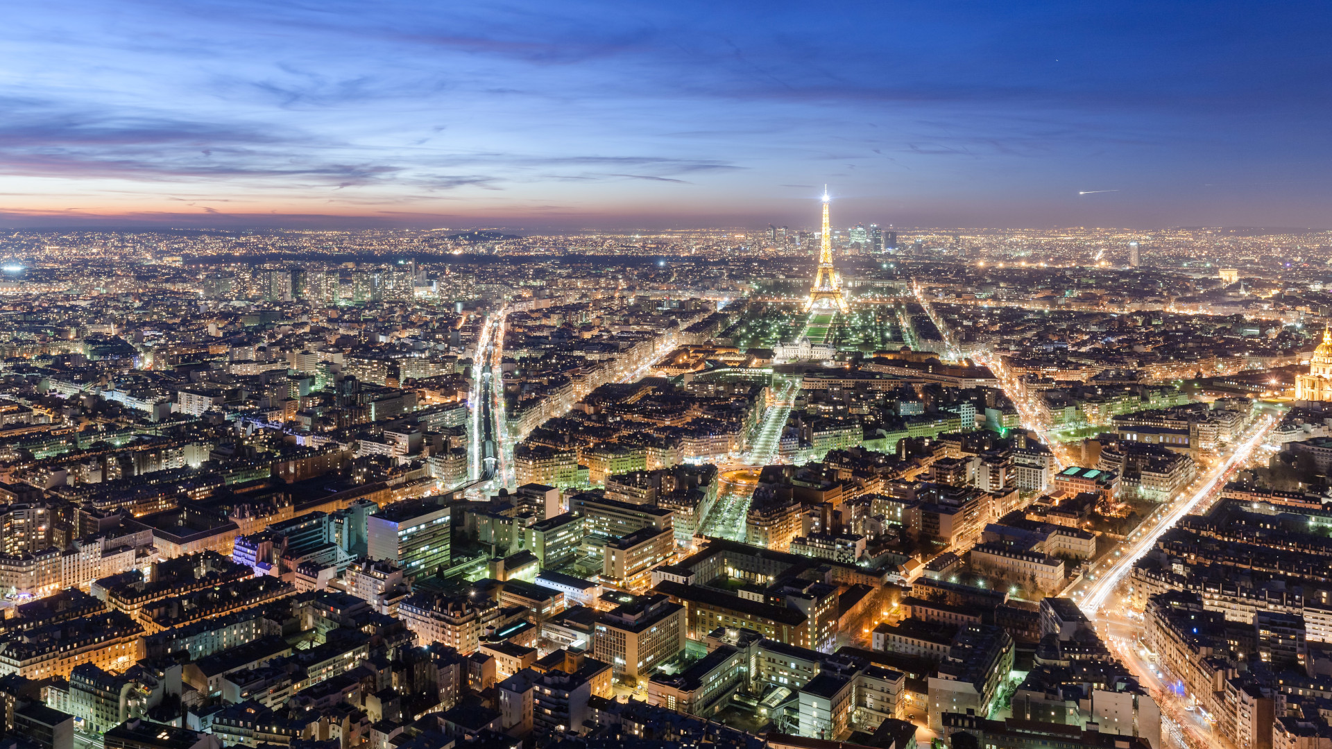 Widok z lotu ptaka na Paryż o zmroku. W jednej trzeciej obrazu widoczna linia horyzontu z wąskim fragmentem czerwonego nieba rozświetlonymi ostatnimi promieniami słońca. Wyraźnie widoczne światła domów oraz latarni wzdłuż ulic, a także Wieża Eiffla.