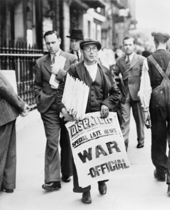 Mężczyzna na ulicy z plikiem gazet pod pachą, trzymający w drugiej ręce rozwinięty biały plakat z napisem: „DISPATCH / SPECIAL LATE NEWS / WAR – OFFICIAL”. W tle przechodnie.