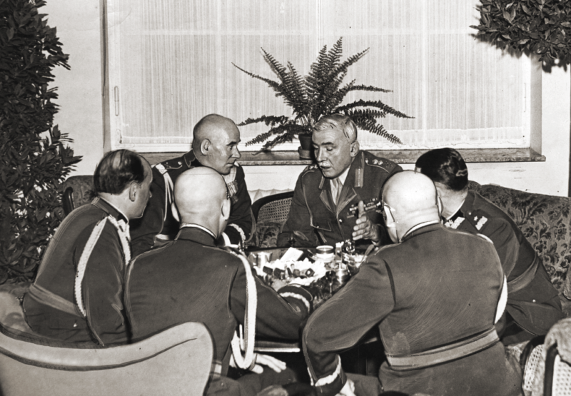 Sześciu mężczyzn w mundurach wojskowych rozmawia dookoła niewielkiego okrągłego stolika.