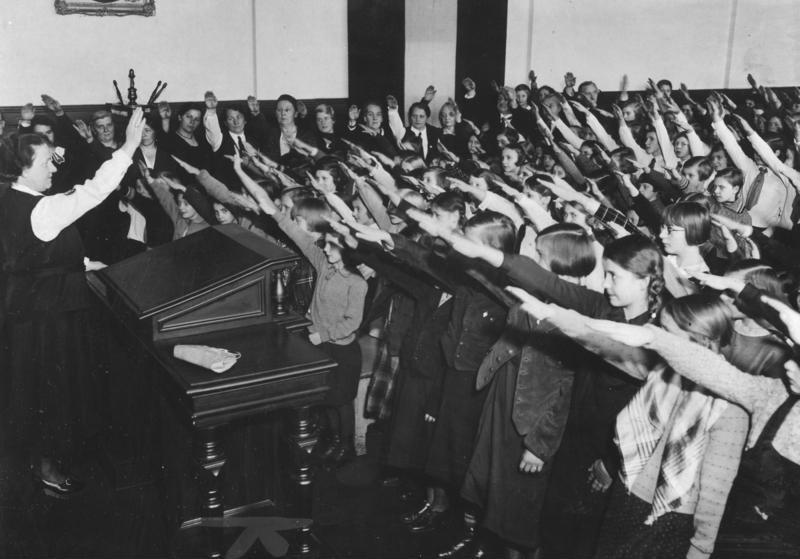 Dziewczynki i chłopcy w wieku ok. 10 i 11 lat stoją przed nauczycielką na katedrze. Wszyscy wznoszą prawe ręce w geście Heil Hitler.