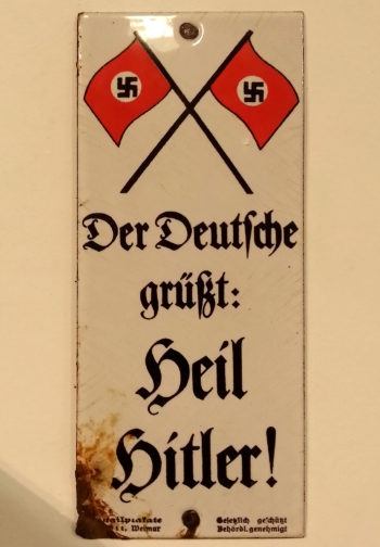 Wąska, pionowa, emaliowana tabliczka przymocowana do ściany dwoma gwoździami (u góry i u dołu). U góry dwie skrzyżowane nazistowskie flagi (czerwone ze swastyką po środku), a niżej zapisano pismem gotyckim po niemiecku: „Niemiec pozdrawia: Heil Hitler!”