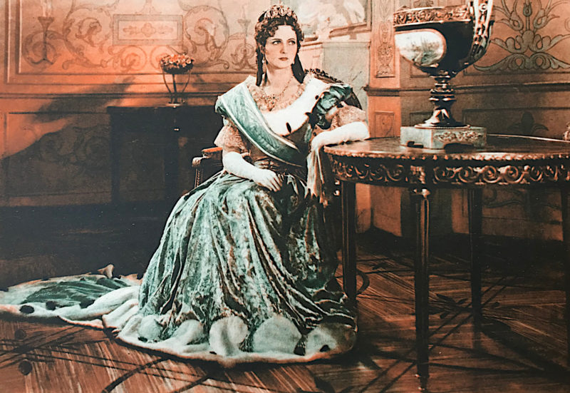 Jadwiga Smosarska siedząca na krześle przy okrągłym stoliku w pałacowym wnętrzu. Ma na sobie bardzo bogatą suknię oraz diadem.