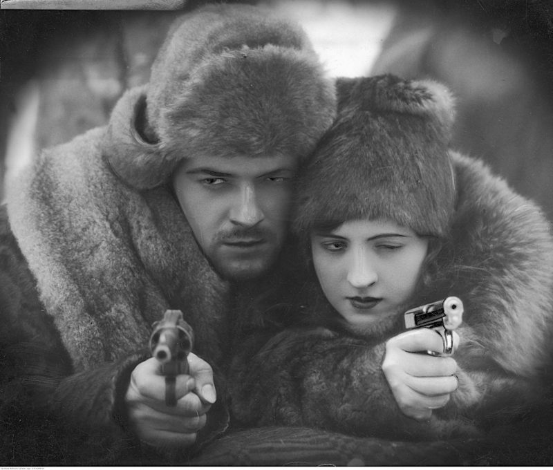 Adam Bronisz i Jadwiga Smosarska w ciepłych futrzanych czapkach i korzuchach, stojący lub siedzący twarzą przy twarzy. Oboje trzymają pistolety i mierzą do celu.