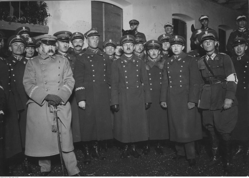 Marszałek Józef Piłsudski wśród oficerów 1 Pułku Szwoleżerów, biorących udział w zawodach jeździeckich Pułku. Marszałek Józef Piłsudski (z lewej, w jasnym płaszczu) w otoczeniu oficerów. Widoczni m.in.: ppłk Jan Głogowski (na prawo za Marszałkiem), gen. Gustaw Orlicz-Dreszer (na prawo od ppłk. Głogowskiego), rtm. Wacław Calewski (2. z prawej na 1. planie), marzec 1926 r. (fot. Narodowe Archiwum Cyfrowe, sygn. 3/1/0/7/1376/1, domena publiczna)