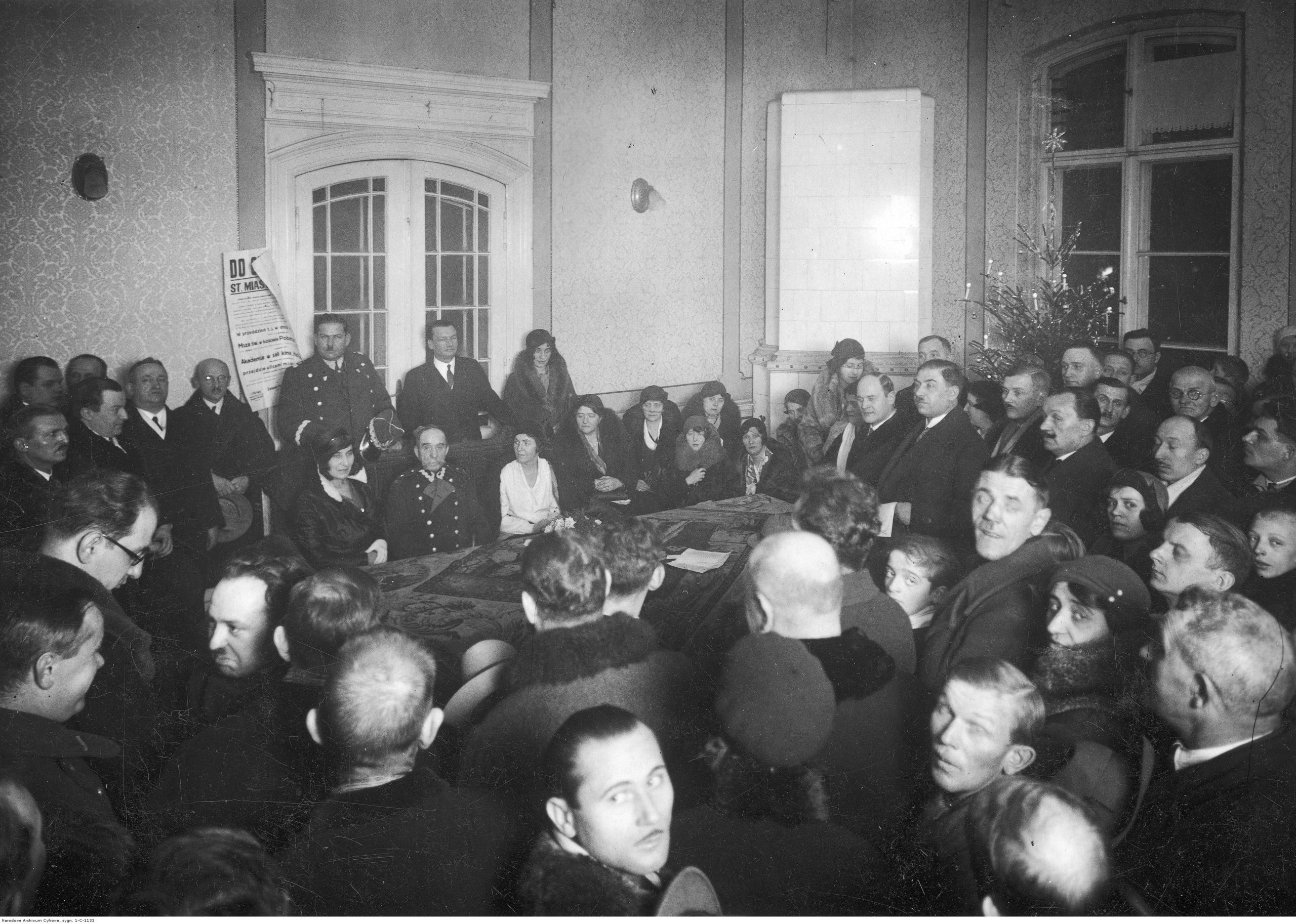 Gwiazdka dla bezrobotnych członków organizacji kombatanckich w Poznaniu. Widoczni m.in.: generał Oswald Frank (stoi bliżej środka z lewej strony przy drzwiach w mundurze), wojewoda poznański Roger Raczyński (stoi na prawo od gen. O. Franka), żona prezesa Zarządu Okręgowego Związku Legionistów profesora Antoniego Jakubskiego (siedzi przy stole w jasnej sukience), żona generała Stanisława Taczaka (siedzi przy stole na lewo od M. Michalskiego), weteran powstania styczniowego Michał Michalski (siedzi przy stole obok kobiety w jasnej sukience); grudzień 1932 r. (fot. Narodowe Archiwum Cyfrowe, sygn. 3/1/0/13/1133/1, domena publiczna)